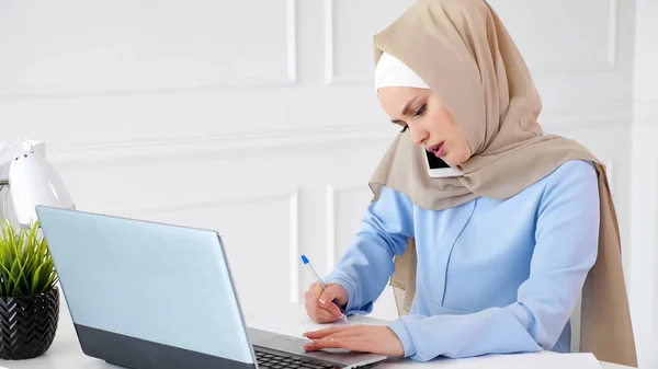 Muslim woman is talking with client on mobile phone, making paper notes and looking at laptop screen.