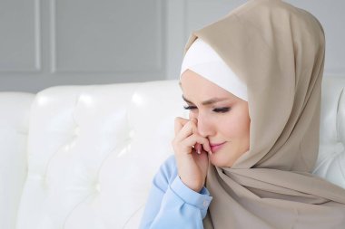 Sad crying muslim woman in hijab is sitting on the sofa at home. clipart