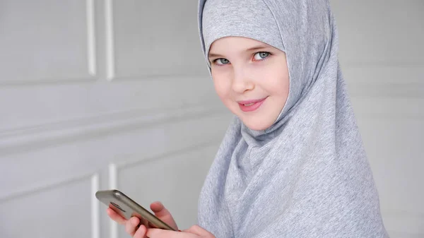 Young muslim teen girl in hijab is playing smartphone, shyly looking at camera and smiling. — ストック写真