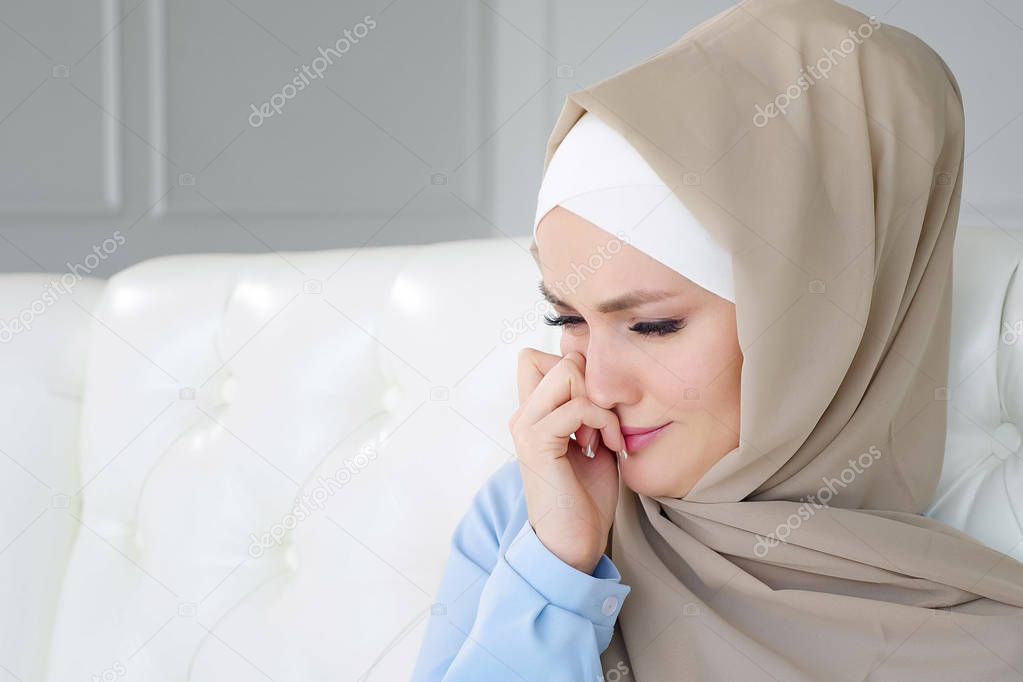 Sad crying muslim woman in hijab is sitting on the sofa at home.