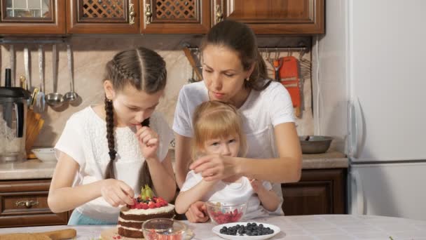 Familie, moeder met twee kleine dochters zijn versieren verjaardagstaart met bessen samen in de keuken thuis. — Stockvideo