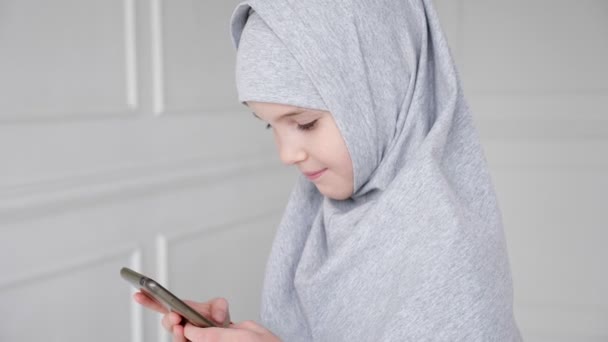 Young muslim teen girl in hijab is playing smartphone, shyly looking at camera and smiling. — ストック動画