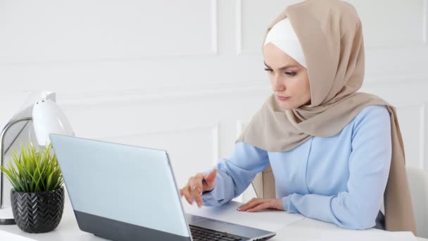 Junge muslimische Studentin im Hijab schreibt und zeichnet ihre Hausaufgaben mit Laptop auf Papier. — Stockvideo