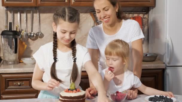 Familie, moeder met twee kleine dochters zijn versieren verjaardagstaart met bessen samen in de keuken thuis. — Stockvideo