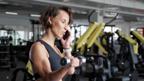 Volwassen vrouw is het maken van biceps oefening met dumbbells in de sportschool en spreken telefoon. — Stockvideo