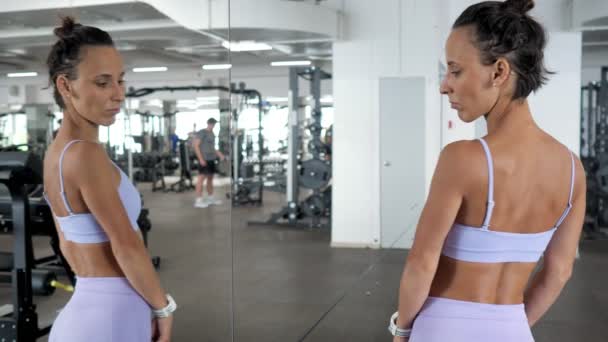 Mujer está mirando el espejo en su hermoso cuerpo deportivo después del entrenamiento en el gimnasio . — Vídeo de stock