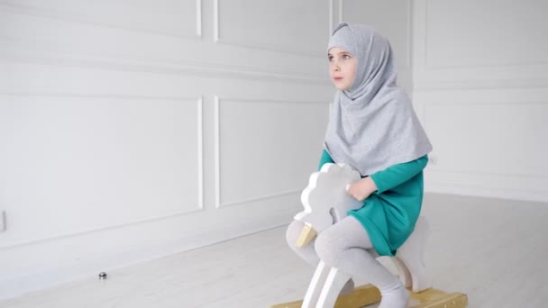 Musulmana adolescente chica en hijab y vestido es jugar a montar en juguete caballo mecedora en su habitación . — Vídeo de stock