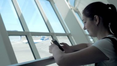 Genç kadın havaalanında uçuş bekliyor cep telefonu ekranına bakıyor.