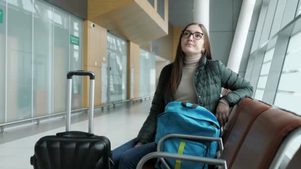 Joven mujer cansada en gafas está esperando su vuelo en el aeropuerto sentado en la sala . — Vídeos de Stock