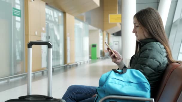 Женщина в аэропорту ждет свой рейс, что-то глядя на смартфон . — стоковое видео