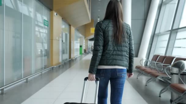 Mujer viajero está caminando en la moderna terminal del aeropuerto que lleva la maleta, vista trasera . — Vídeos de Stock