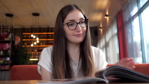 Glimlachend mooi meisje in glazen uitziende menu in café. — Stockvideo