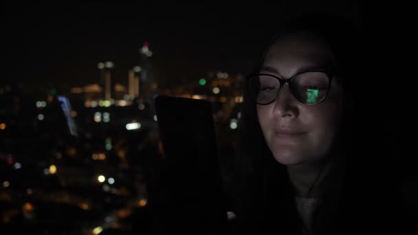 Ospalá žena v noci na telefonu převracuje stránky u okna s výhledem na město.