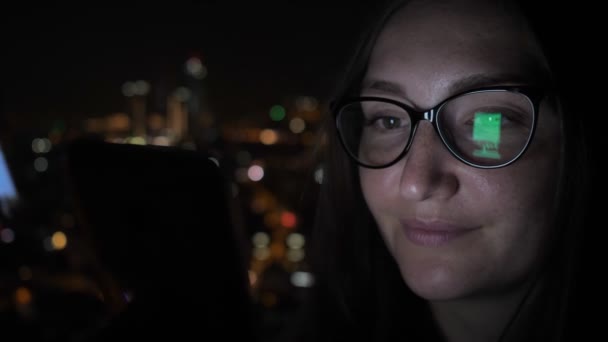 Mujer sonriente está mirando el teléfono inteligente cerca de la ventana con vista a la ciudad, vista lateral . — Vídeo de stock