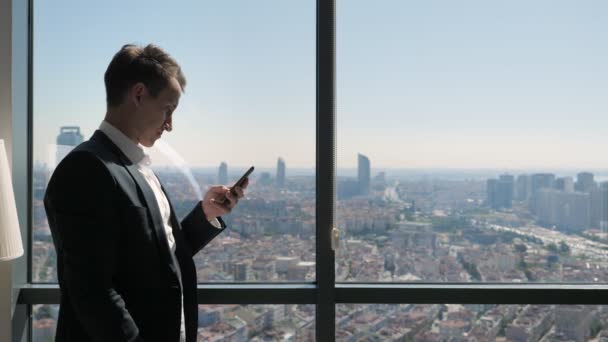 Młody biznesmen w biurze przegląda telefon w pobliżu okna z panoramicznym widokiem na miasto. — Wideo stockowe