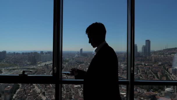 Silueta de joven hombre de negocios en la oficina está buscando teléfono cerca de la ventana con vista panorámica de la ciudad . — Vídeos de Stock