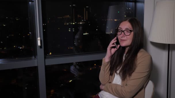 Жінка розмовляє телефоном, сидячи біля вікна з панорамним видом на місто вночі . — стокове відео