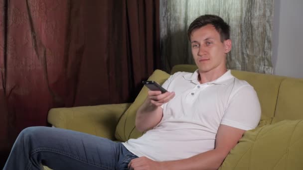 Jovem homem rindo em camiseta branca está sentado no sofá assistindo TV — Vídeo de Stock