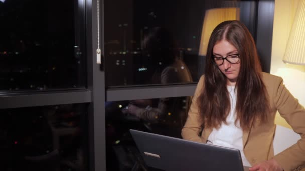 Vrouw in glazen werkt overuren op computer in kantoor met panoramisch uitzicht op de stad. — Stockvideo