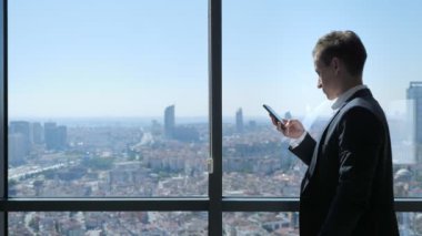 Ofistegenç işadamı panoramik şehir manzaralı pencerenin yanında telefon tarama.