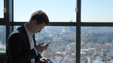 İşadamı şehir manzaralı pencerenin yanında oturan telefon ve dizüstü bilgisayar ile çalışıyor.