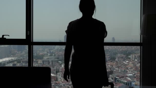 Silhouette of young girl traveler with suitcase is entering in hotel room with panoramic city view. — Stock Video