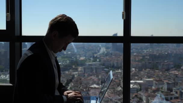 Ofiice worker in suit is typing on laptop sitting near the window with panoramic city view. — Stock Video