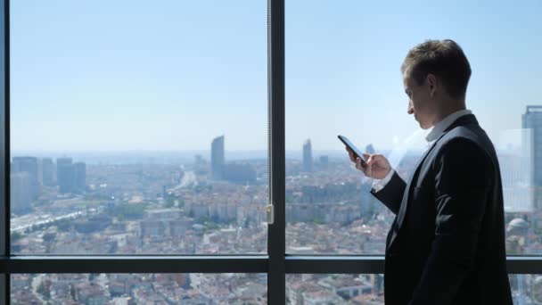 Młody biznesmen w biurze przegląda telefon w pobliżu okna z panoramicznym widokiem na miasto. — Wideo stockowe