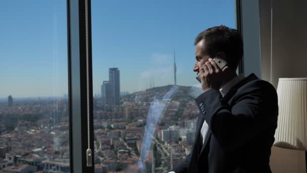 Ofis kıyafeti genç işadamı panoramik şehir manzaralı pencerenin yanında telefon konuşuyor. — Stok video