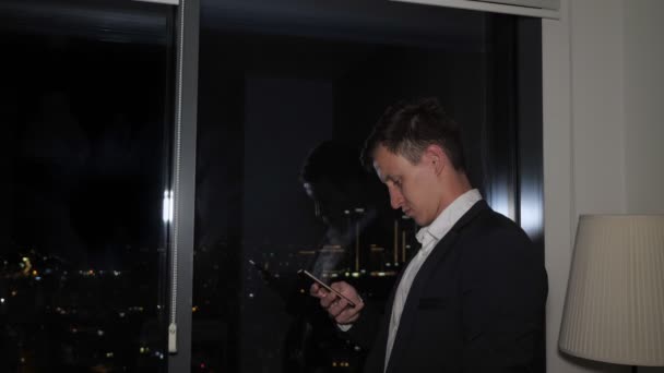 Jonge zakenman in Office Suit is browsen smartphone in de buurt van het raam met uitzicht op de stad. — Stockvideo
