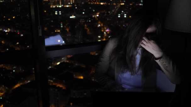 Tired sleepy woman is working on her computer at night near the window with cityscape. — Stock Video