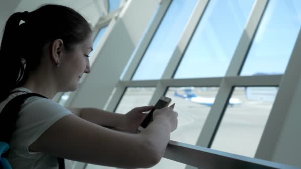 Mujer joven está mirando a la pantalla del teléfono móvil esperando su vuelo en el aeropuerto . — Vídeos de Stock