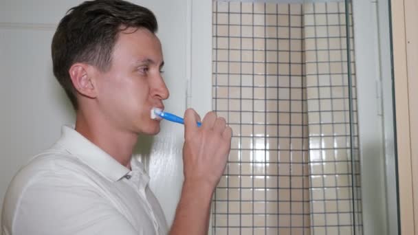 Jovem está escovando os dentes no banheiro de manhã olhando para o espelho . — Vídeo de Stock