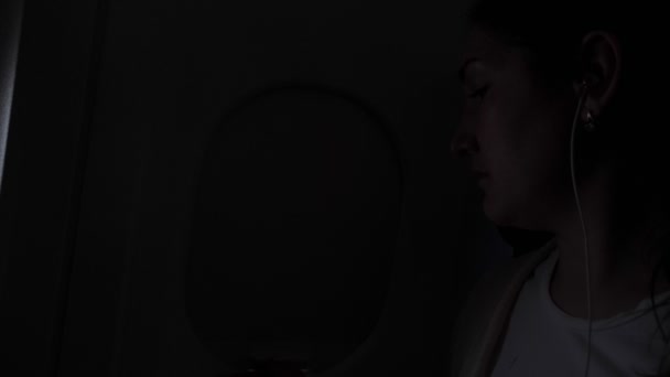 Mulher ouve música em fones de ouvido durante o voo no escuro e abre a janela . — Vídeo de Stock