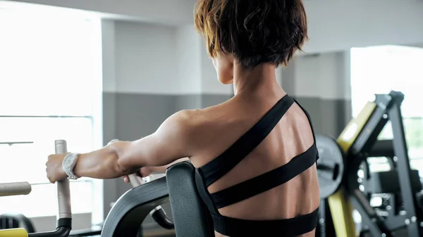Mulher está fazendo exercícios para os músculos da coluna vertebral na máquina de remo, visão traseira . — Fotografia de Stock