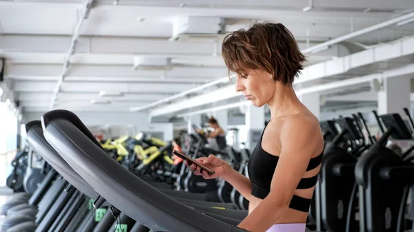 Mulher desportiva está treinando em esteira na academia e olhando no smartphone . — Fotografia de Stock