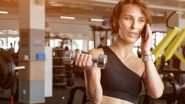 Dospělá žena dělá biceps s činkami v tělocvičně a mluví telefonem. — Stock fotografie