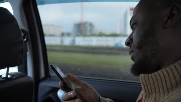 Retrato de hombre negro navegando teléfono inteligente está montando un coche en día lluvioso en la ciudad . — Vídeos de Stock