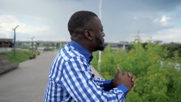 Hombre negro se para en el paseo marítimo de la ciudad cerca de valla en el parque y admira la vista de la ciudad . — Vídeos de Stock