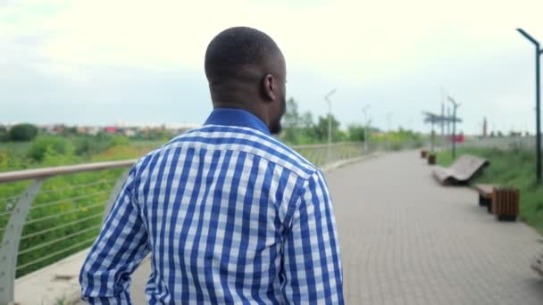 Jeune homme afro-américain marche dans le parc de la ville tourne la tête et sourit à la caméra . — Video