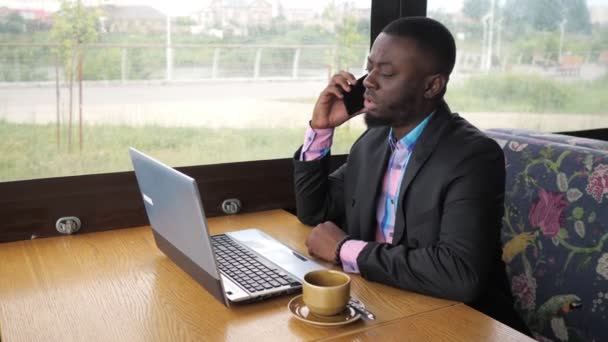 黑人商人在笔记本电脑上工作,并调用智能手机坐在咖啡与咖啡. — 图库视频影像