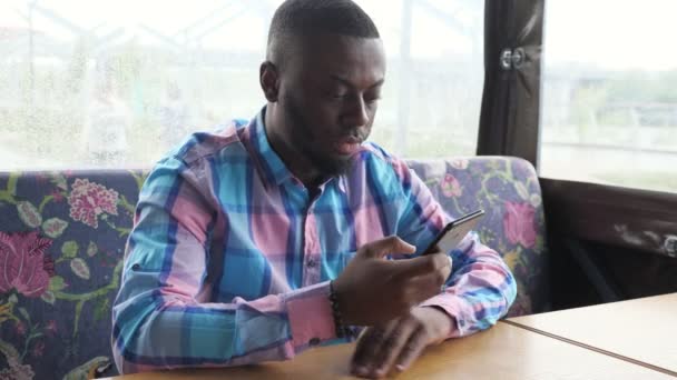 アフロアメリカ人の男は、彼の注文を待ってカフェに座ってスマートフォンを閲覧しています. — ストック動画