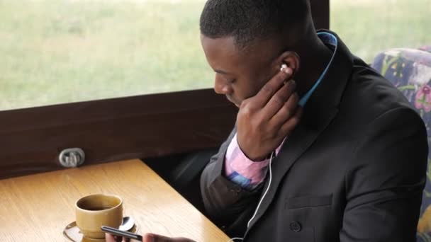 Homme d'affaires noir écoute de la musique dans les écouteurs assis dans le café et boit du café . — Video