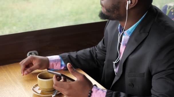 Homem preto está ouvindo música em fones de ouvido navegando smartphone sentado no café . — Vídeo de Stock