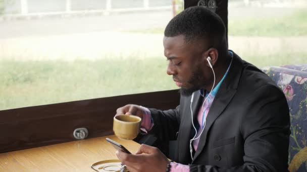 카페에 앉아 스마트폰을 검색하는 이어폰에서 음악을 듣는 흑인. — 비디오