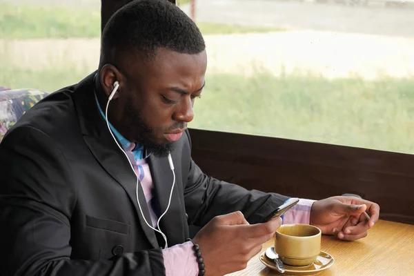 Negro hombre está escuchando música en los auriculares de navegación smartphone sentado en la cafetería . — Foto de Stock