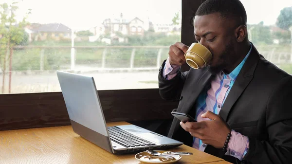 L'uomo d'affari nero lavora su computer portatile con smartphone in caffè e bevande caffè . — Foto Stock