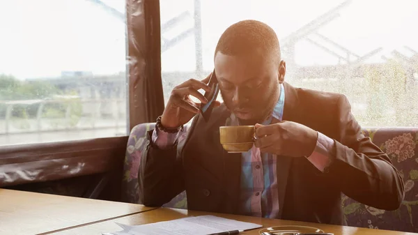 Afro americano uomo d'affari sta chiamando su smartphone e bere caffè in caffè . — Foto Stock