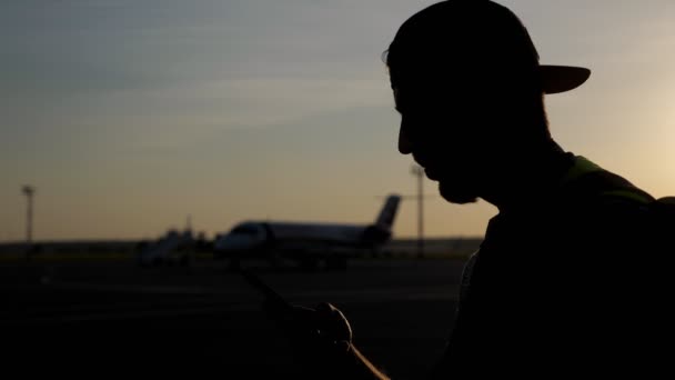 Неузнаваемый человек просматривает мобильный телефон на аэродроме с самолетом на заднем плане . — стоковое видео
