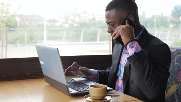 Black businessman works on laptop and calls smartphone sits in cafe with coffee. — Stock Video